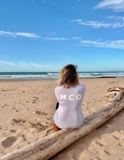Femme en t-shirt gris N’CO regardant l’océan de dos sur la plage