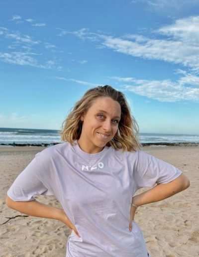 Femme portant un t-shirt gris N’CO de face avec la plage en arrière-plan