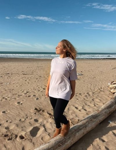 Femme portant un t-shirt gris N’CO face à l’océan sur la plage, les yeux fermés